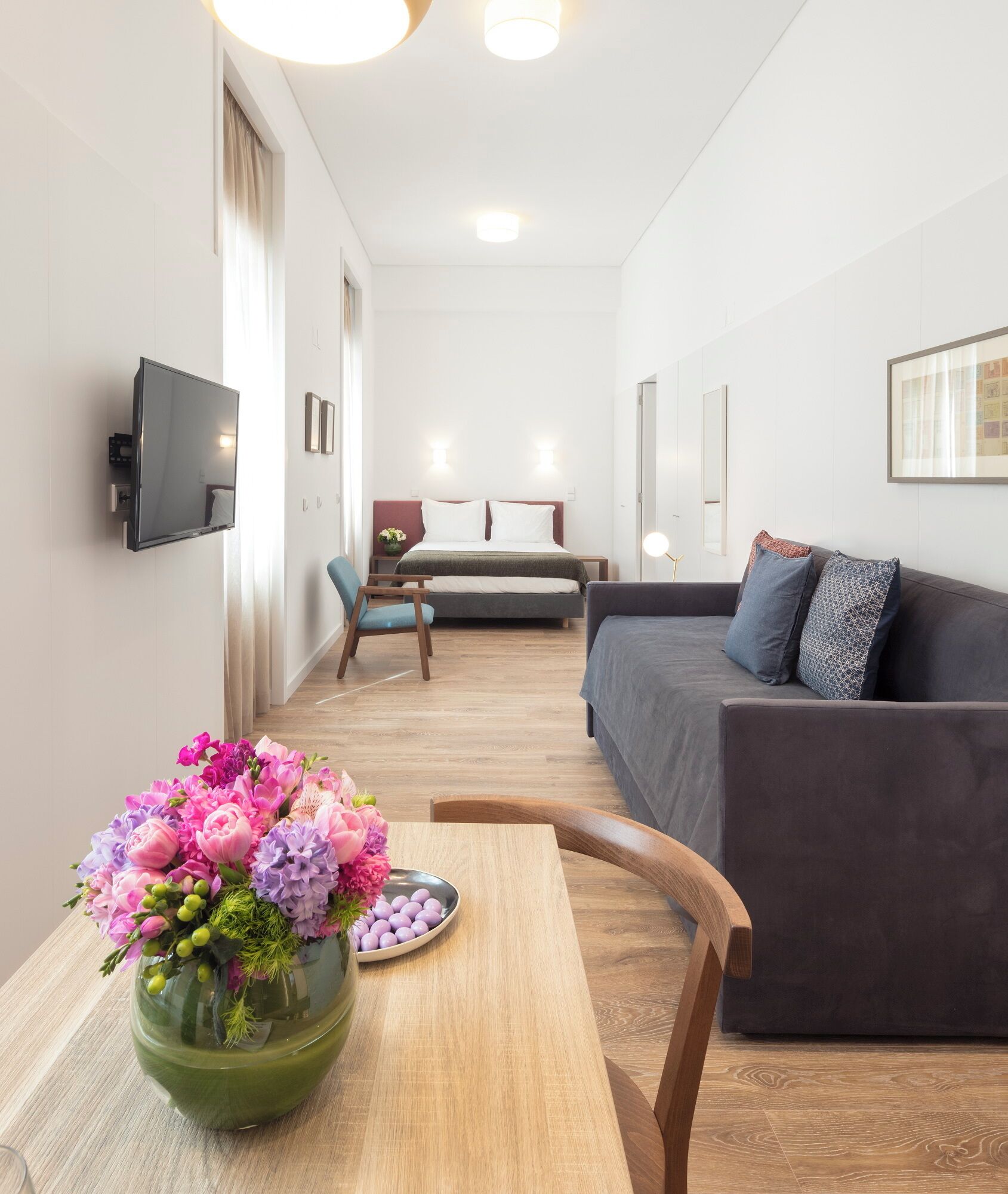 Una sala moderna con un sofá, un televisor montado en la pared, una mesa de comedor con un jarrón de flores y una cama al fondo.