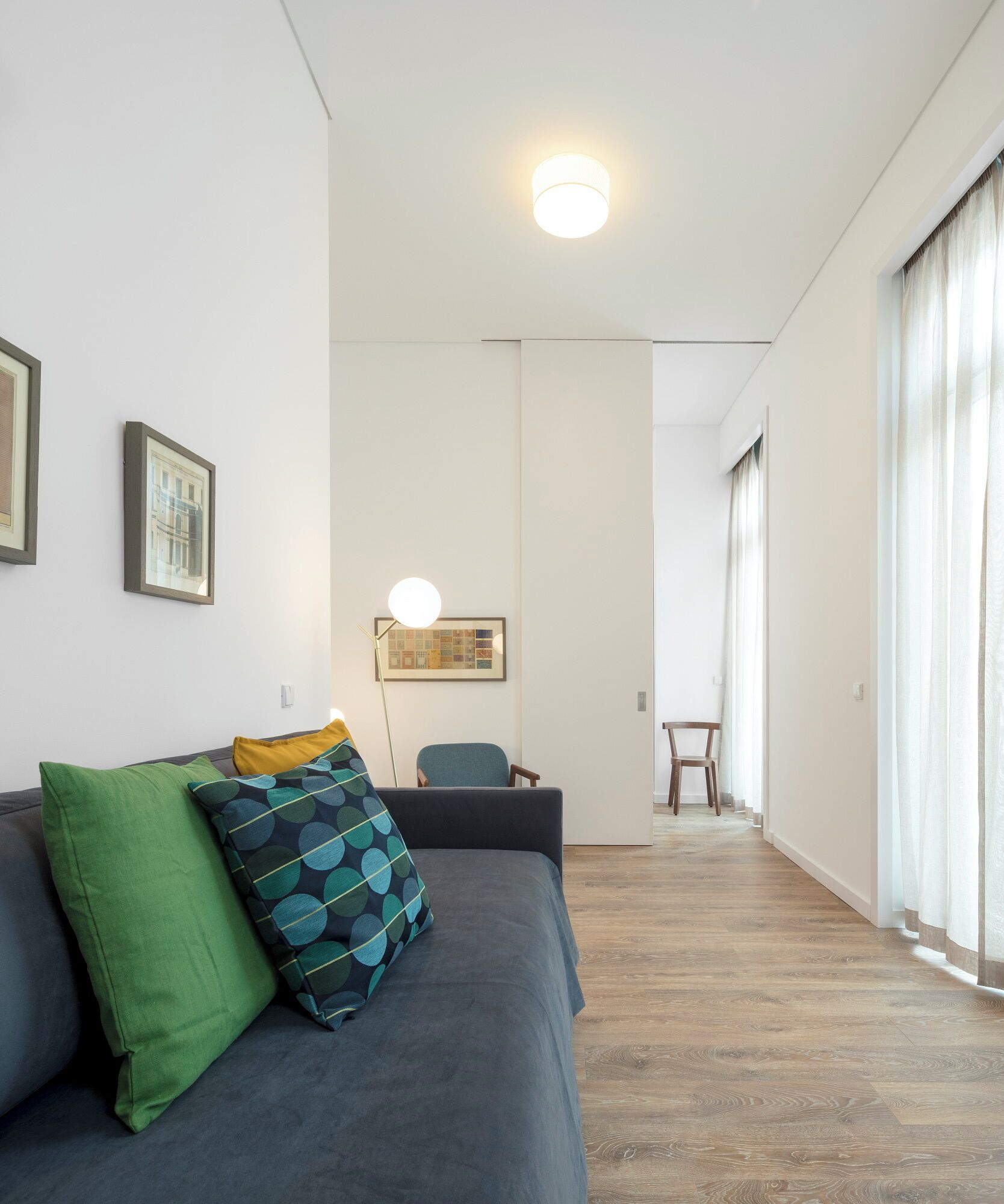 Una sala moderna con un sofá gris, almohadones estampados en verde y azul, una mesita y una silla. Hay un cuadro en la pared y una lámpara de techo.