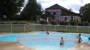 A swimming pool with a white fence, a building labeled "CAMPING" in the background, and a grassy area with trees. - Camping les Craoues (Capvern)
