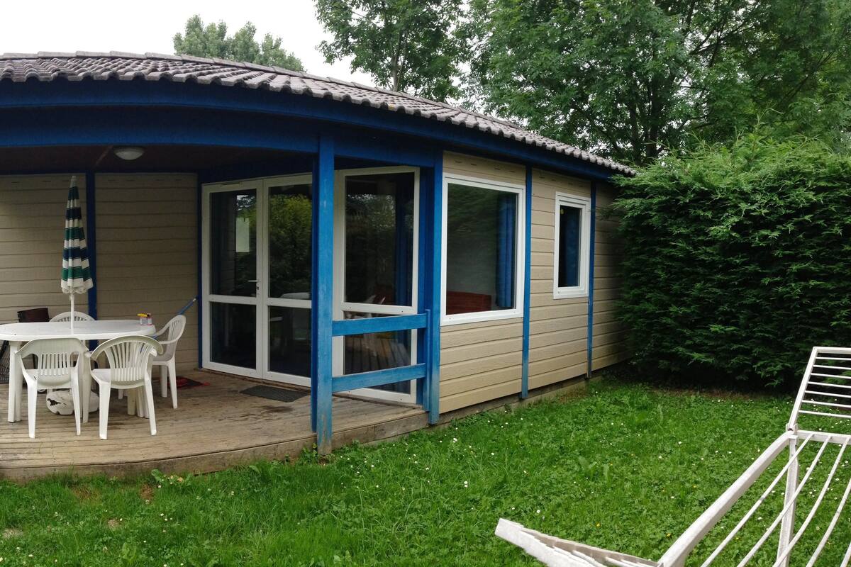 Ein kleines, eingeschossiges Gebäude mit blauem Dach und beigen Wänden, das eine Terrasse mit Tisch und Stühlen sowie umgebende Grünflächen zeigt.