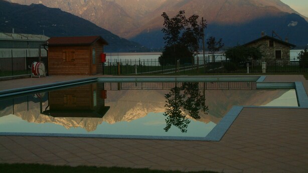 A swimming pool with a mountain backdrop, wooden buildings, and a fenced area. - Hotel Resort Le Vele (Domaso)