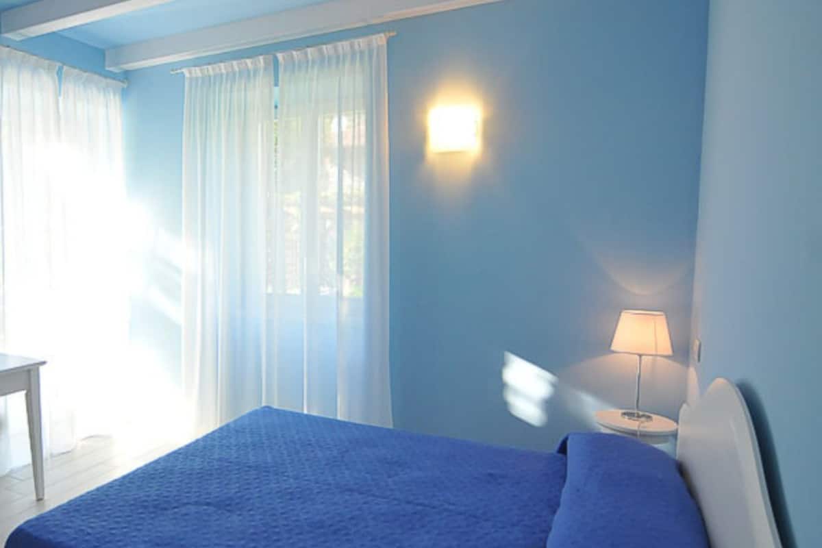 A room with a bed, a nightstand with a lamp, and a window with sheer curtains.