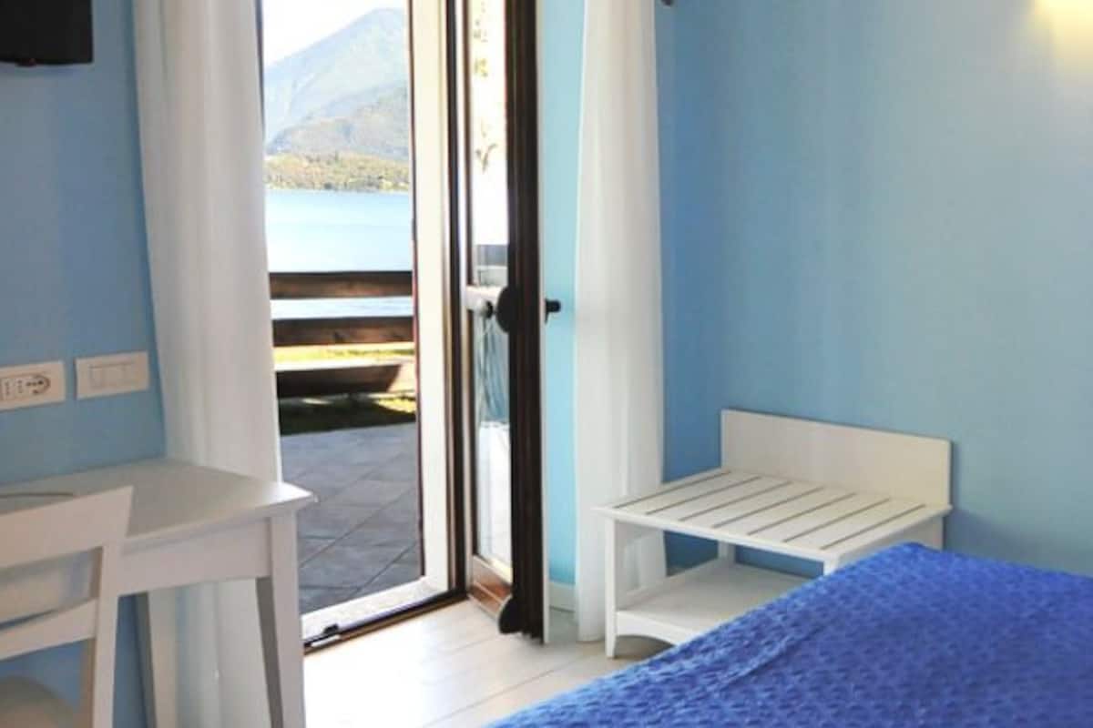A room with a bed, a desk, a chair, a lamp, and a view of a body of water through a door.