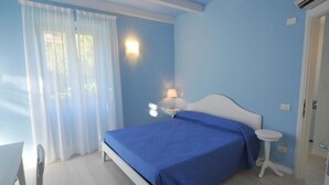 Ein Schlafzimmer mit Bett, Nachttisch mit Lampe, kleinem runden Tisch und einem Fenster mit Vorhängen.
