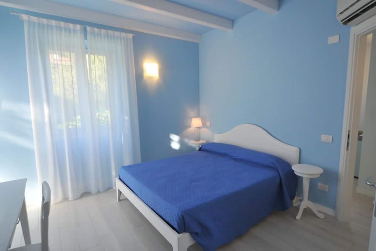 A bedroom with a bed, a bedside table with a lamp, a small round table, and a window with curtains.