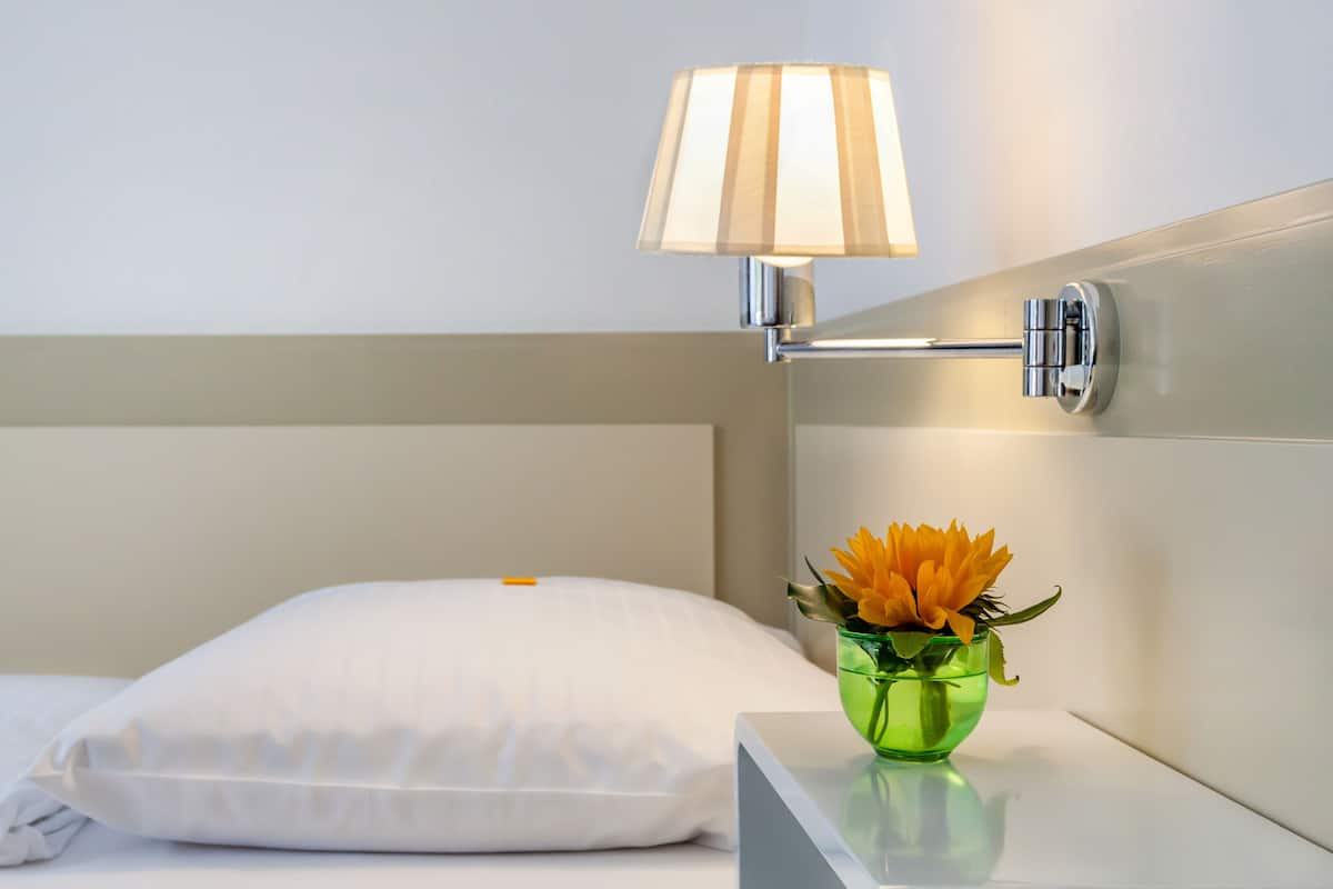 Cama bem arrumada com travesseiro branco e um vaso verde com flores em uma mesa de cabeceira.