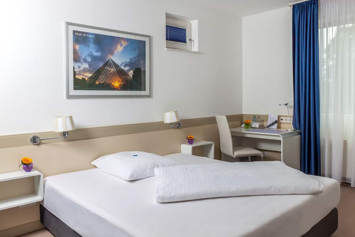 Quarto de hotel com cama, escrivaninha, cadeira, mesa de cabeceira, abajur, moldura de quadro e janela com cortinas.