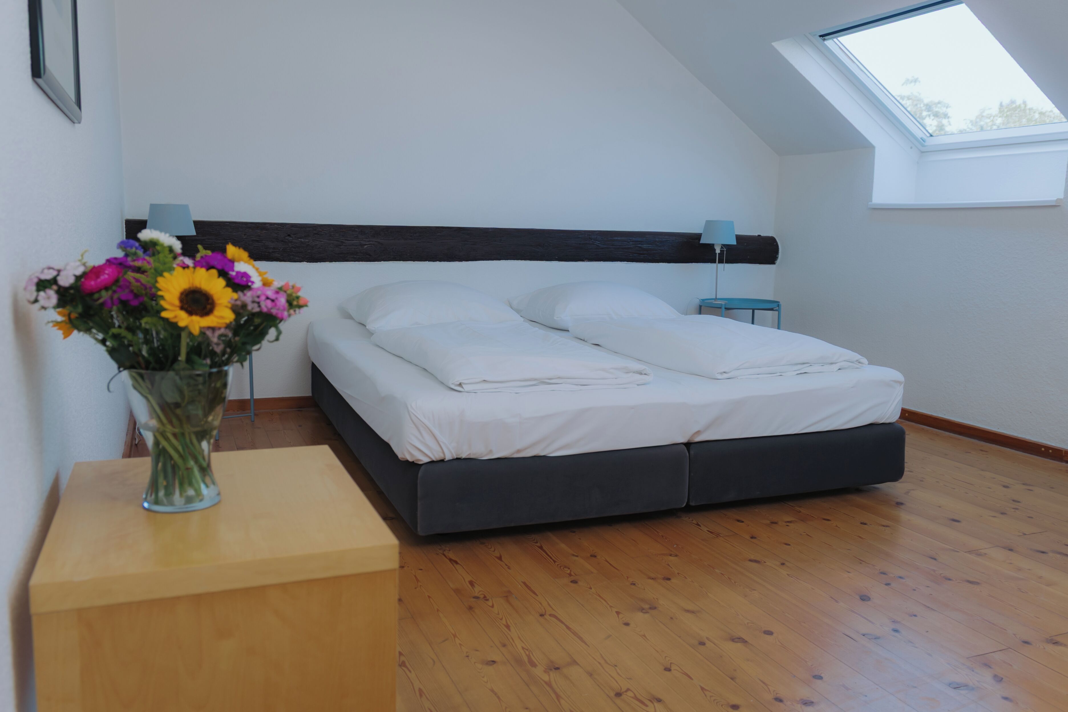 A bedroom with a skylight, a bed with white linens, a bedside table with a vase of flowers, and a wooden floor.
