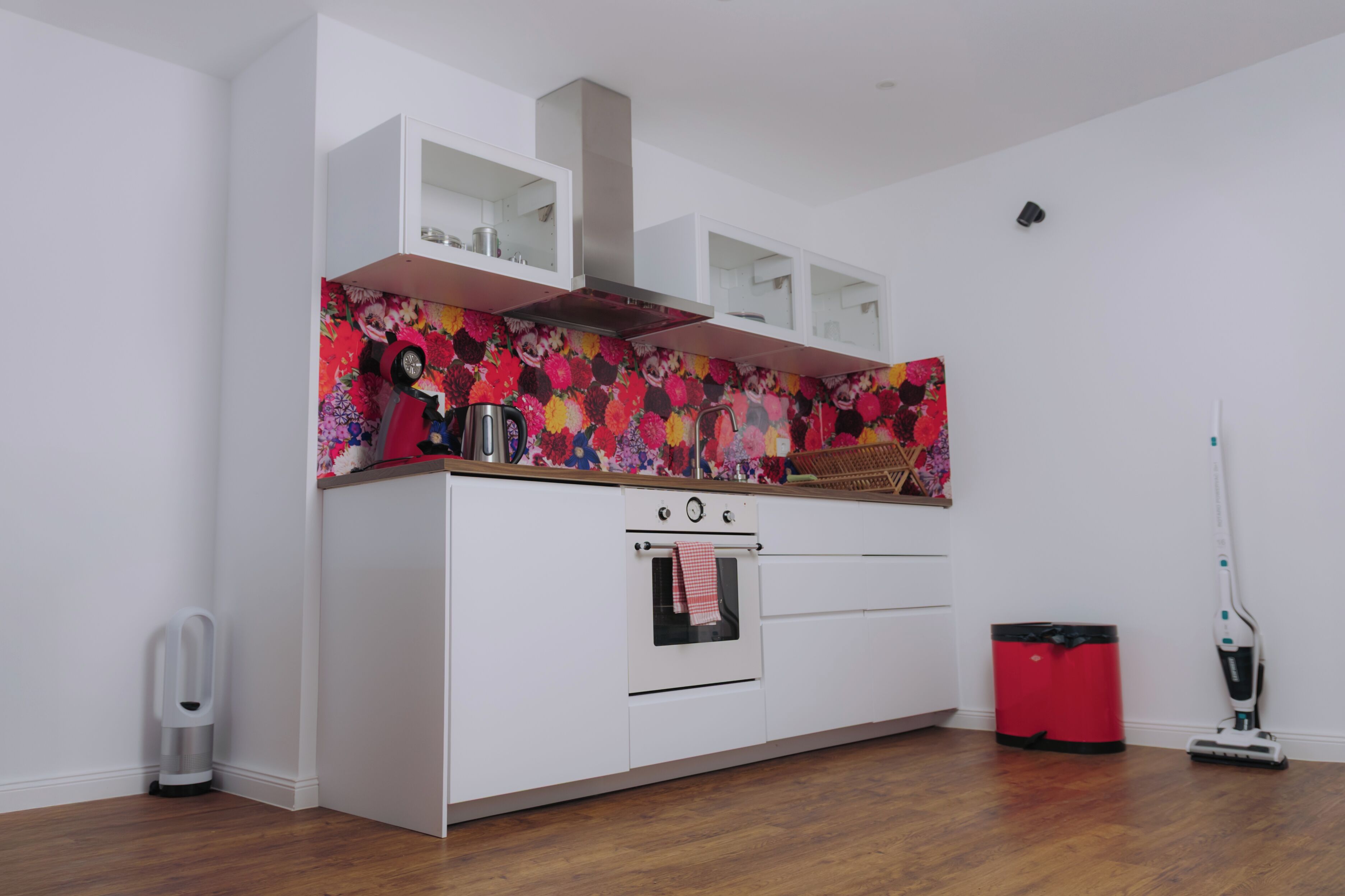 A modern kitchen with white cabinets, a colorful backsplash, a built-in oven, and a red trash bin.