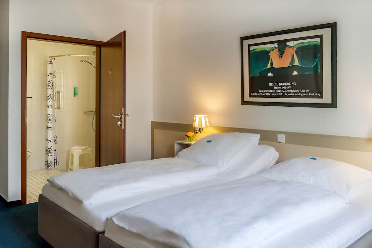 Quarto de hotel com uma cama, mesa de cabeceira com abajur, quadro na parede e área de chuveiro com cortina.