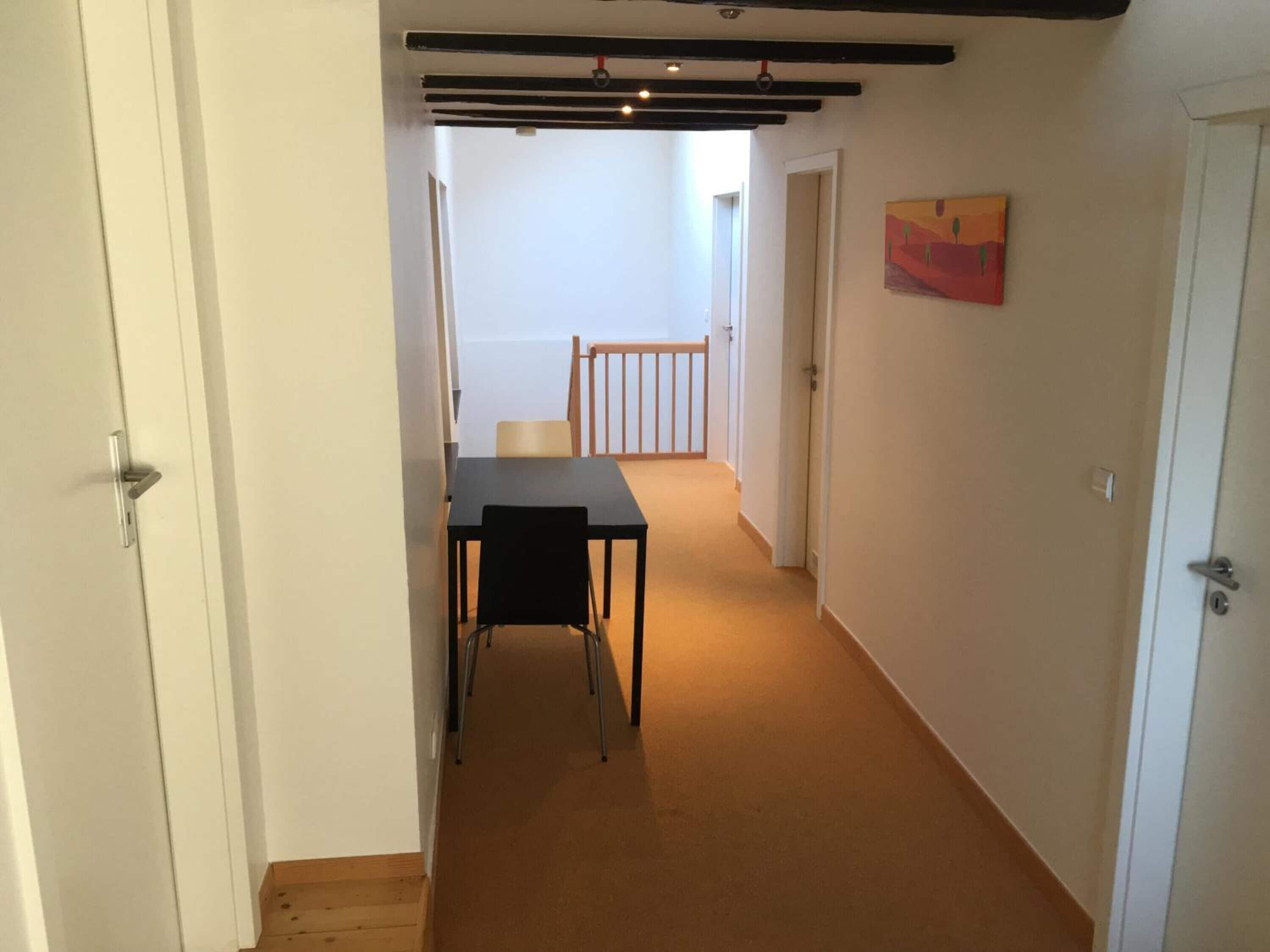 Un passadís amb una taula de menjador i cadires, una cadira de fusta, un quadre a la paret i una escala que condueix a una altra habitació.