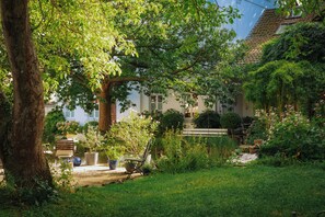 Un jardin avec un banc, des plantes en pot et un bâtiment blanc doté d’un toit recouvert de tuiles.