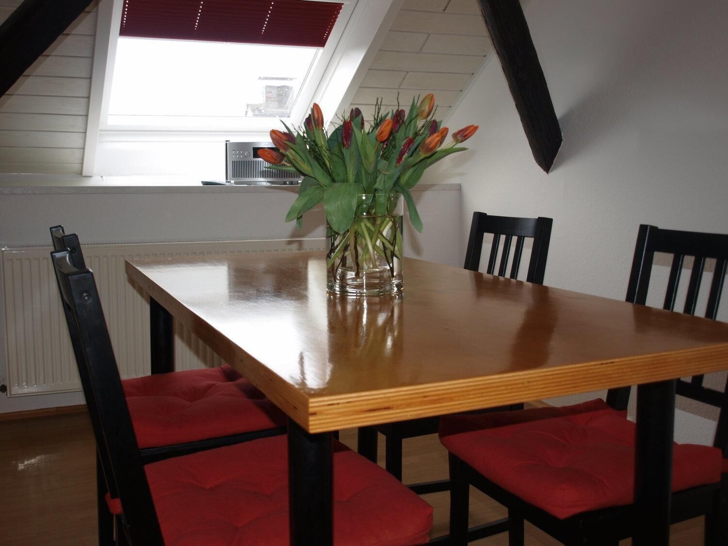 Una mesa de comedor con un jarrón de tulipanes, sillas tapizadas en rojo y una ventana con persianas.