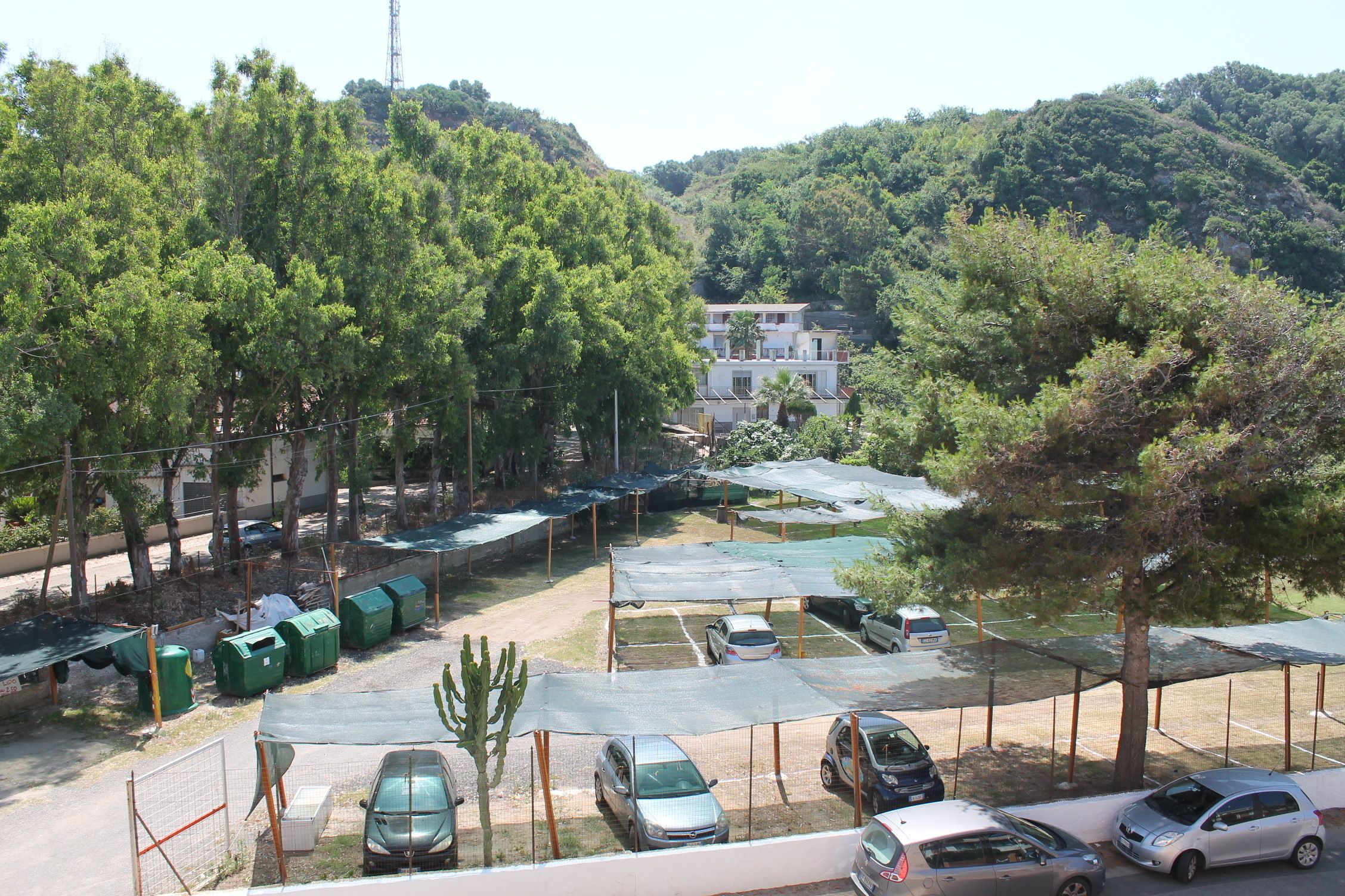 A parking area with multiple cars and covered parking spaces, surrounded by trees and a building in the background.