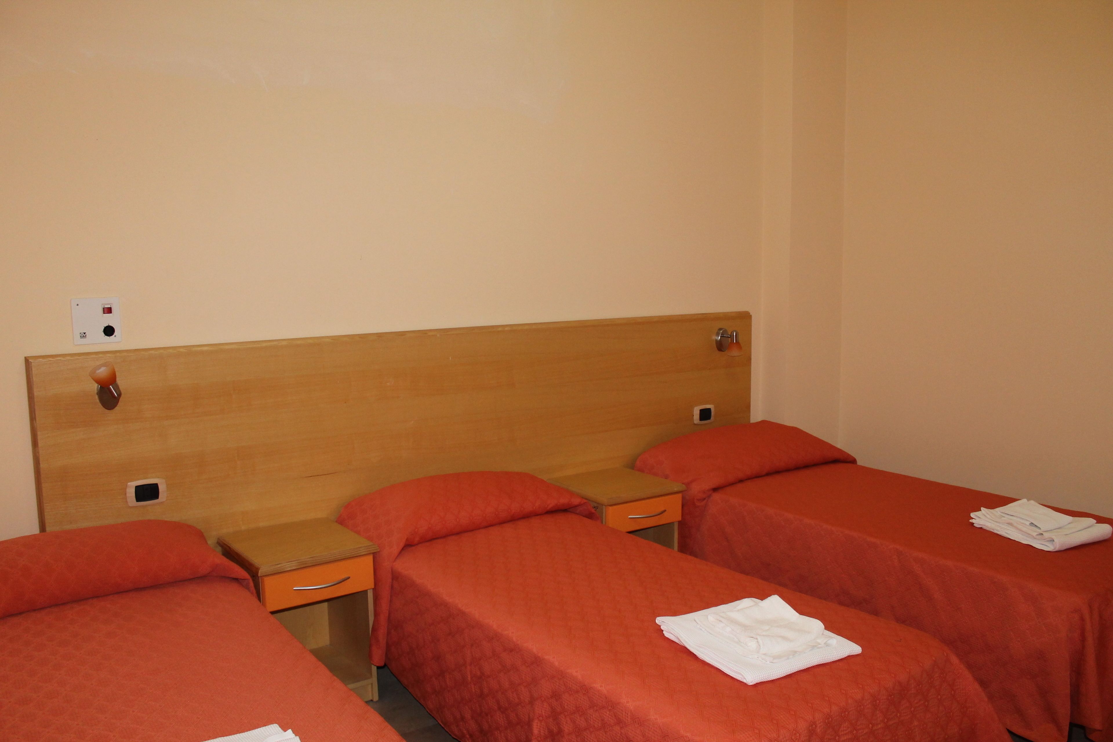 Habitación de hotel con tres camas individuales, cada una con cabecero de madera y ropa de cama naranja. Hay mesitas de noche con cajones y un pequeño estante.