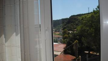 A view through a window with a white curtain, revealing a residential area with houses, trees, and a hill in the background.
