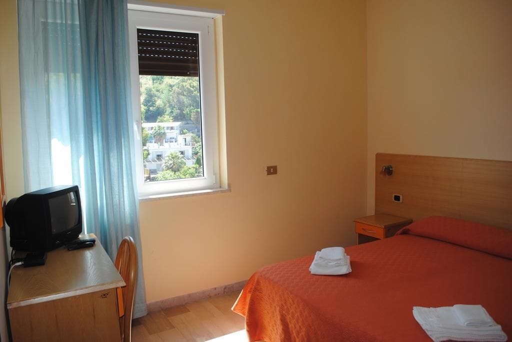 Une chambre d’hôtel avec un lit, un bureau avec une télévision, une fenêtre avec des stores et une vue sur les bâtiments environnants.