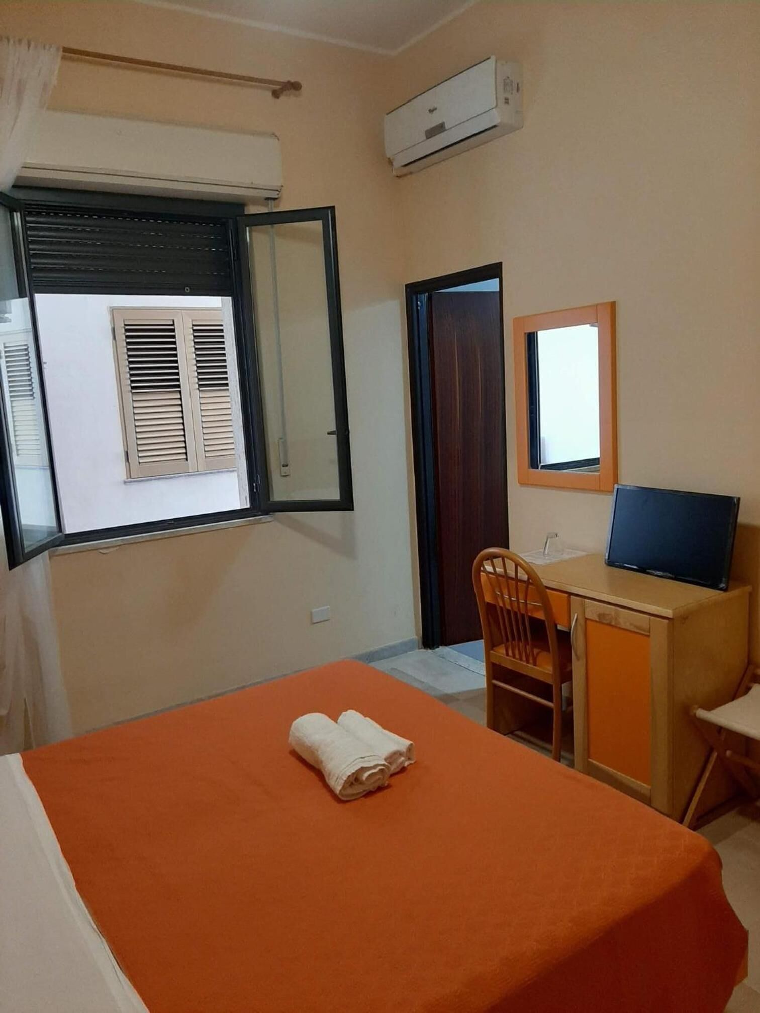 A hotel room with a bed, a desk with a computer, a chair, a window with shutters, and an air conditioning unit.