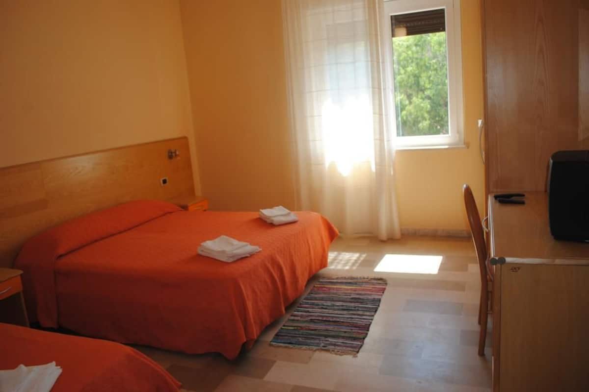 A hotel room with two beds, a wooden headboard, a window with sheer curtains, a small desk, and a television.