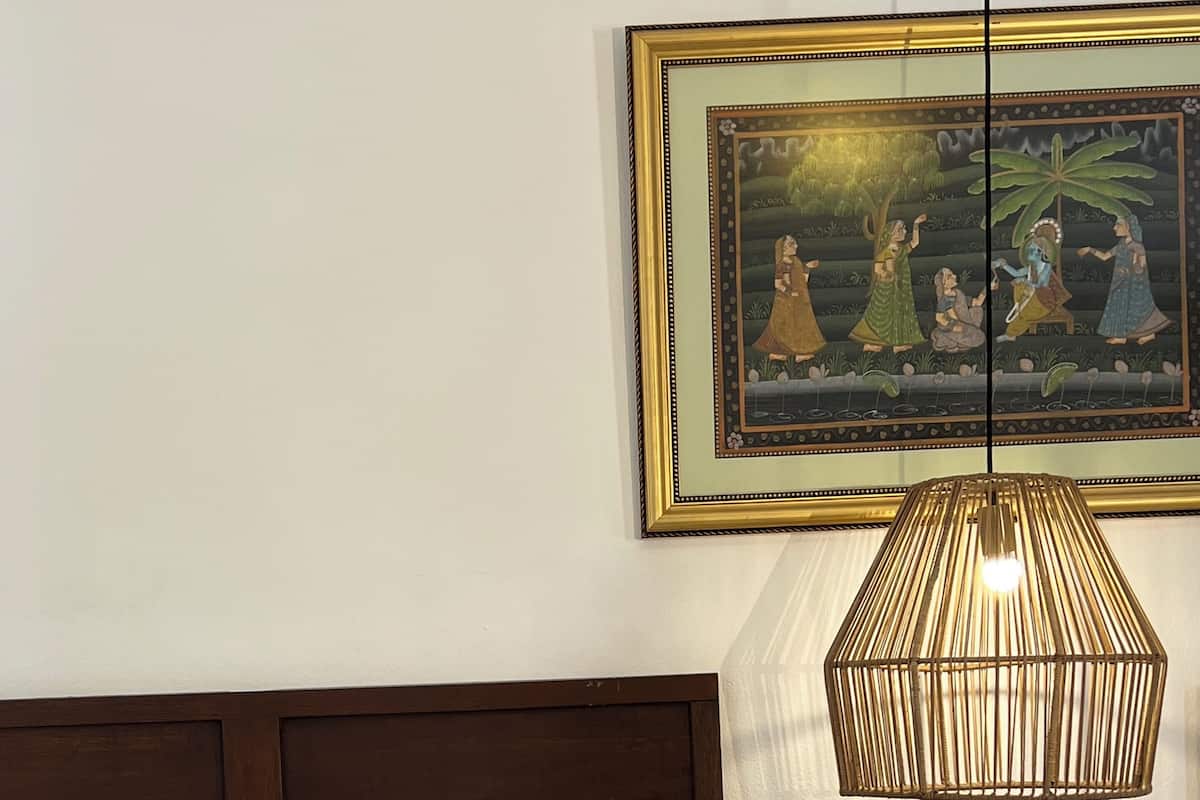 Ein Schlafzimmer mit einem Bett, einer Hängeleuchte, einem kleinen Tisch mit Pflanze und Korb sowie einem gerahmten Gemälde an der Wand.