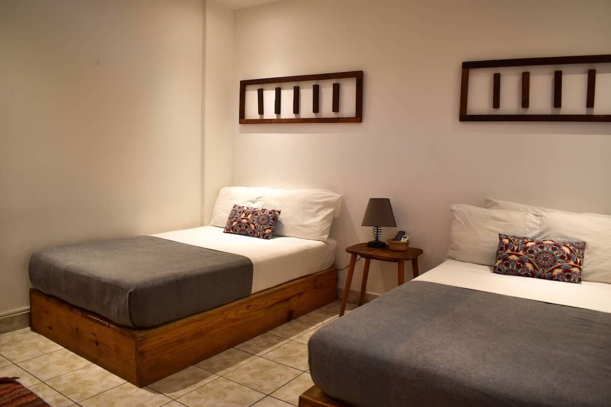 Ein Schlafzimmer mit zwei Betten, jeweils mit grauer Bettdecke und dekorativen Kissen. Ein kleiner Holfnachtschrank mit Lampe und Dekorationselement steht daneben. Der Raum hat einen Fliesenboden und schlichte Wände.