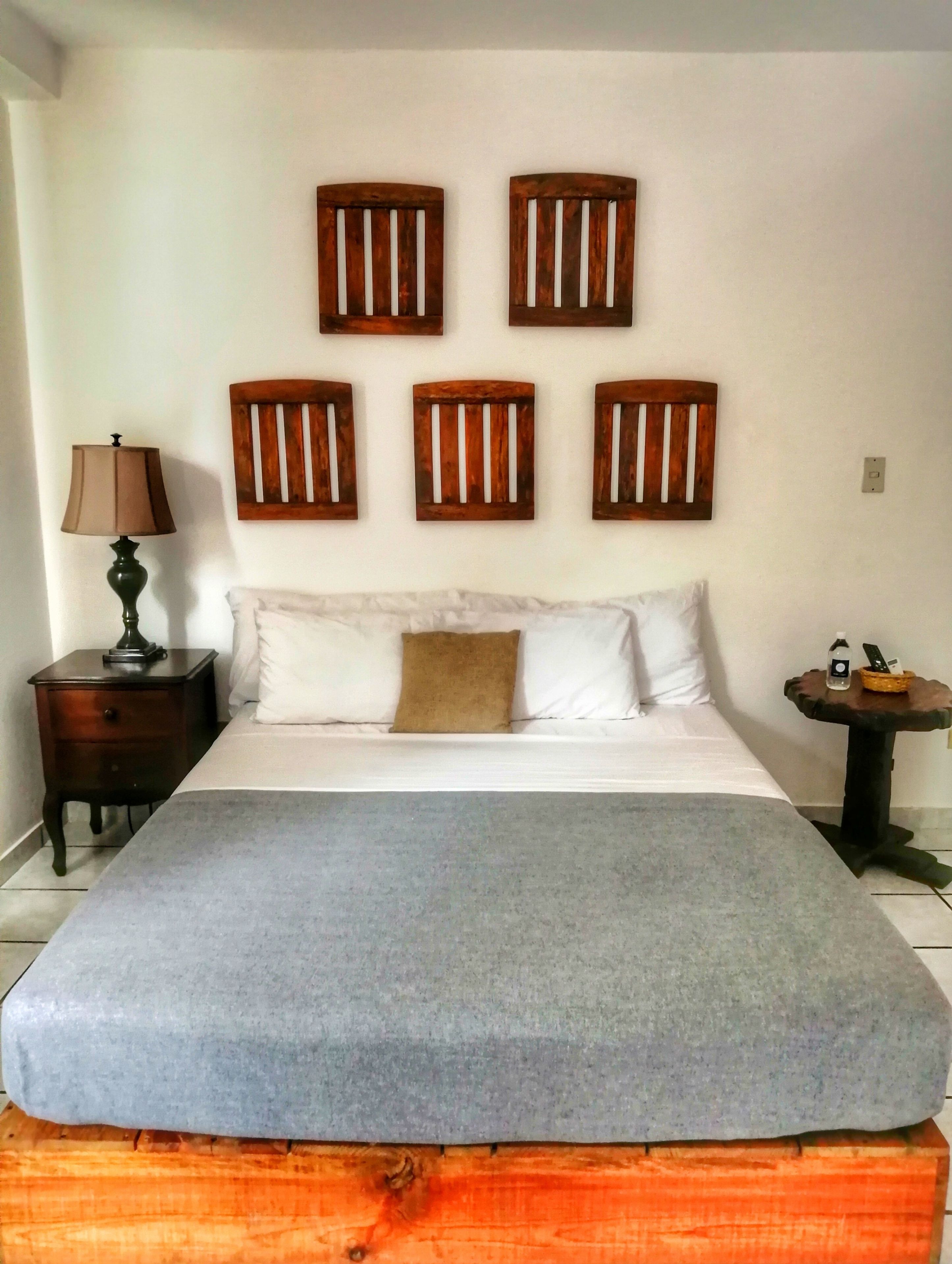 A bedroom with a bed, a nightstand, a lamp, a small table with a phone and a basket, and wooden wall art above the bed.