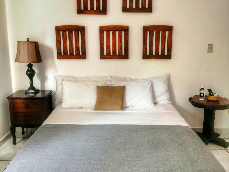 A bedroom with a bed, a nightstand, a lamp, a small table with a phone and a basket, and wooden wall art above the bed.