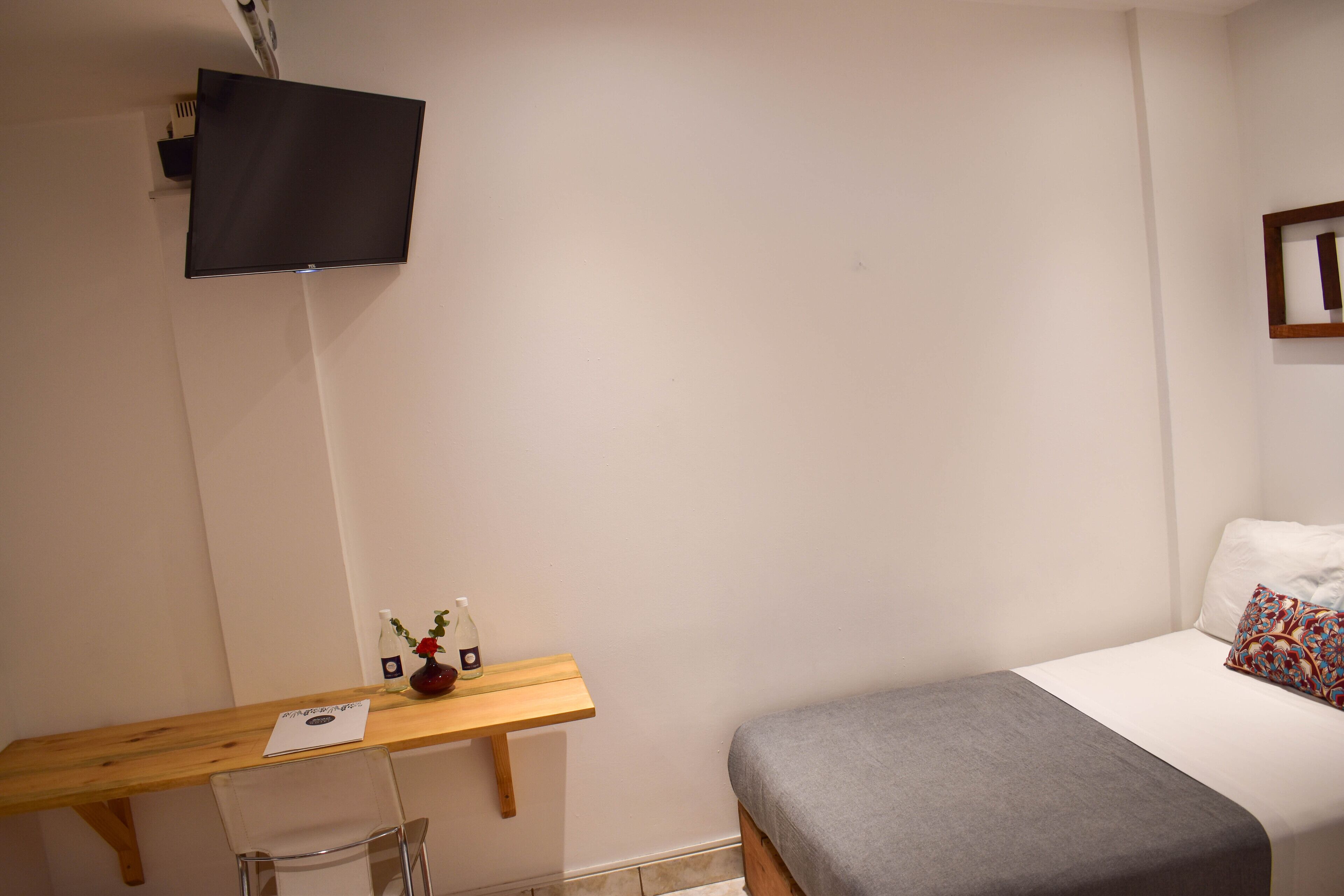 A bedroom with a bed, a bedside table with a vase and a bottle, a wooden desk, and a mounted TV.