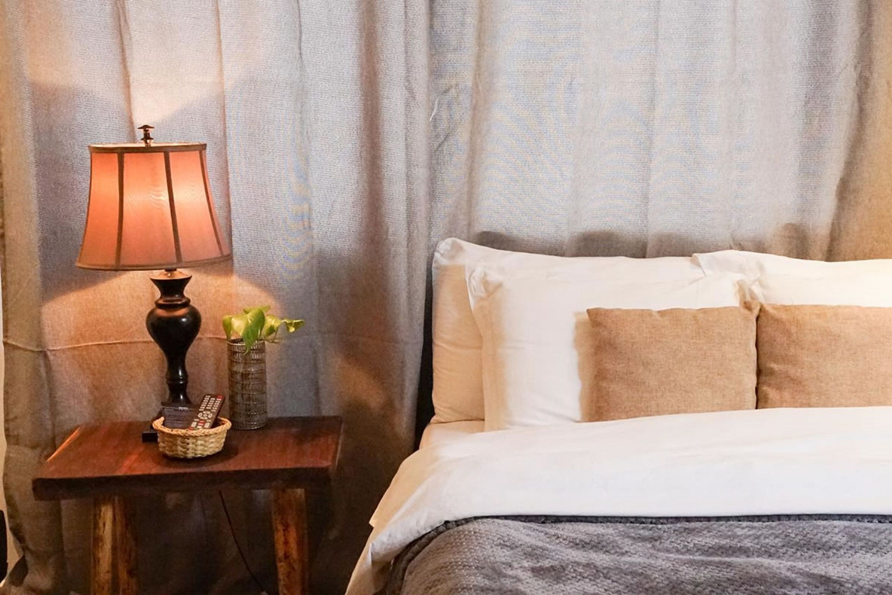 A neatly made bed with white and beige pillows, a bedside lamp, a small vase with a plant, and a basket with remote controls on a wooden nightstand.