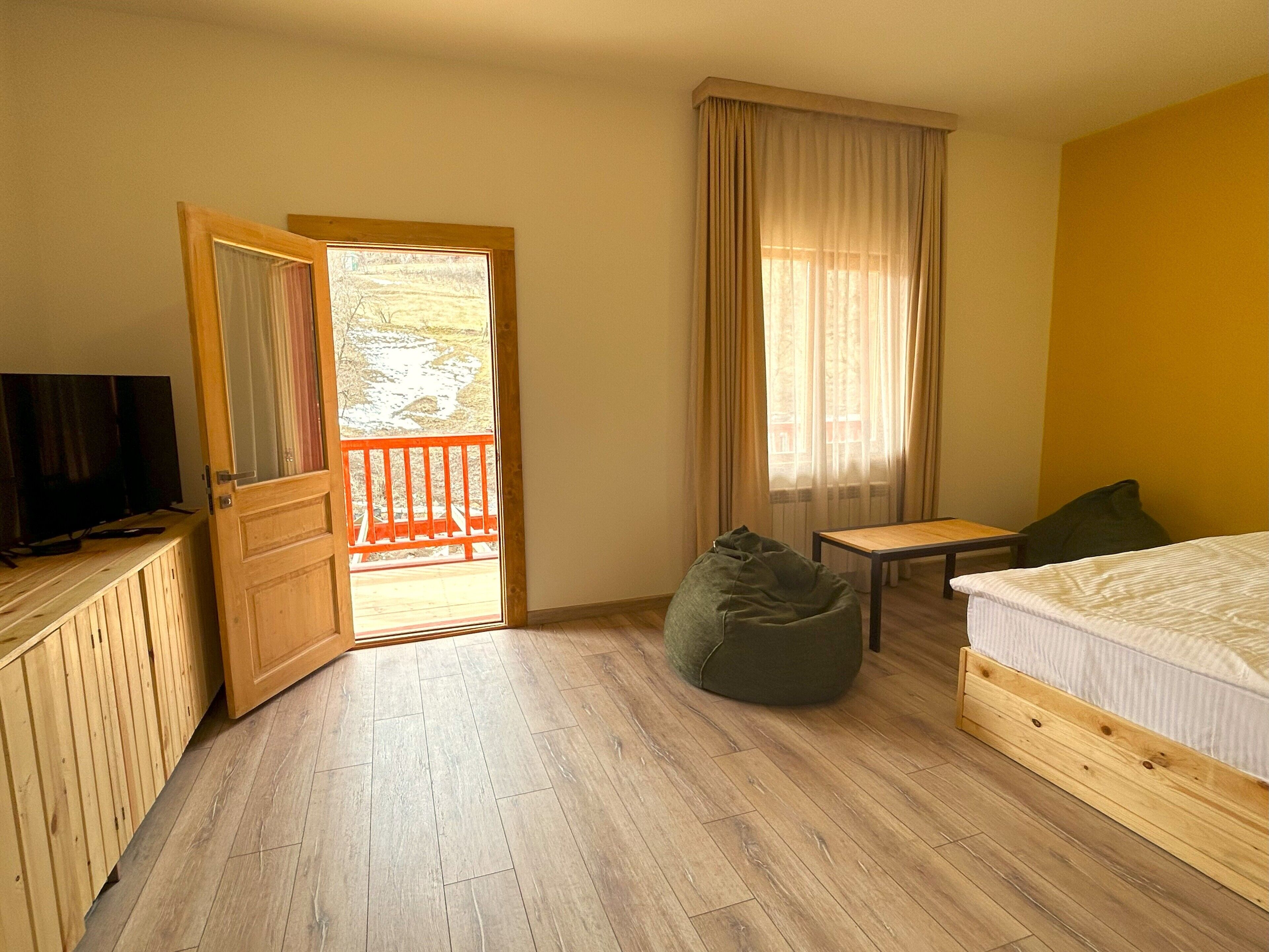 A bedroom with a wooden floor, a bed, a small table, a bean bag, a TV on a wooden cabinet, and a balcony with a red railing.