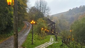 Un sentier rural mène à une maison traditionnelle en bois, dotée d’un toit de chaume, et entourée d’une végétation luxuriante et d’un sol pavé de pierres.