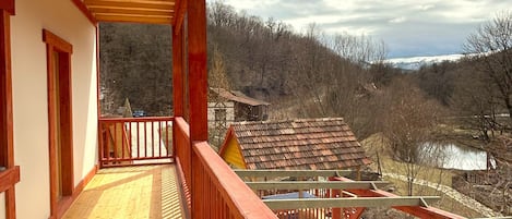 A wooden balcony with a view of a rural landscape, including a pond and buildings in the distance.