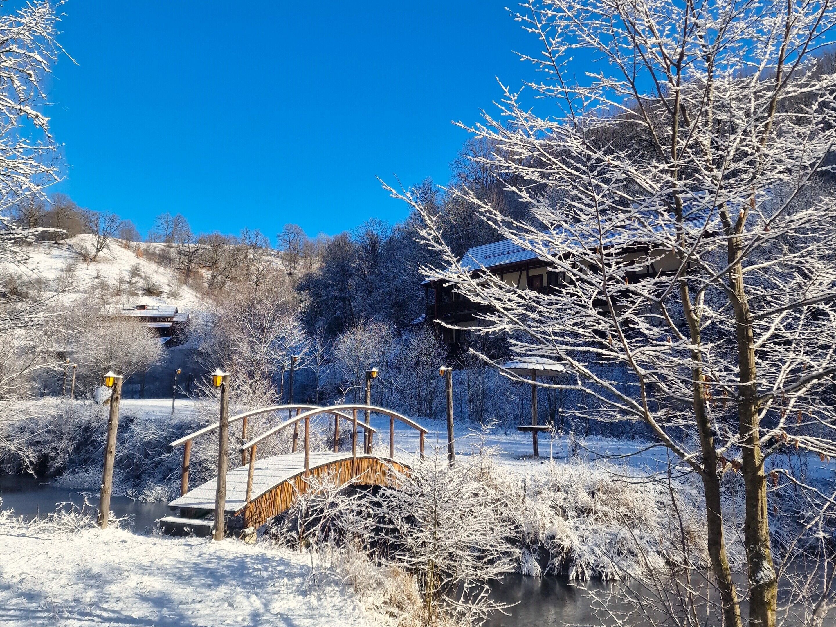 A snow-covered landscape with a wooden bridge, a building with a sloped roof, and a clear blue sky.
