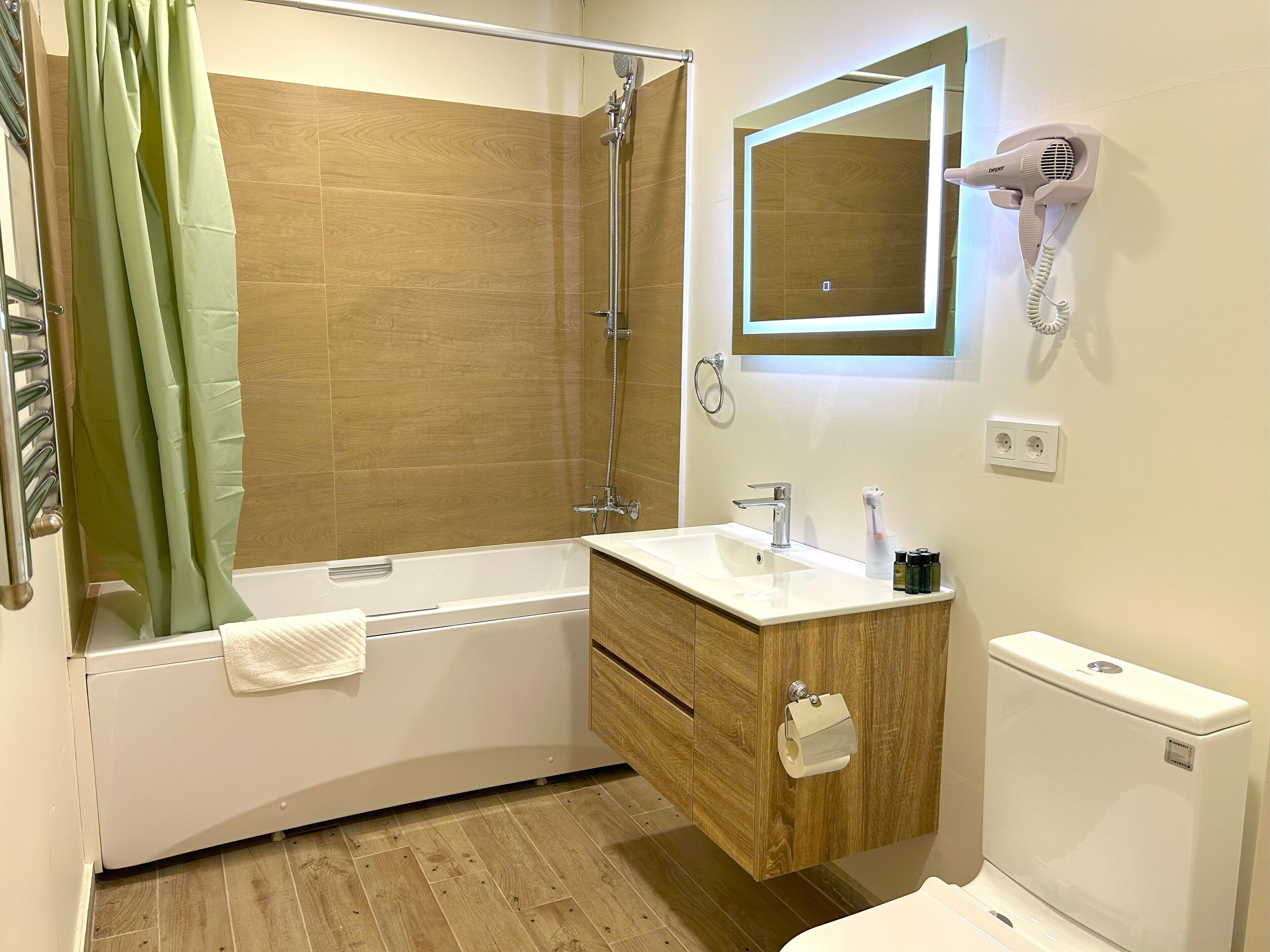 Une salle de bain moderne avec un plancher en bois, une baignoire blanche, un meuble-lavabo en bois, un miroir, un sèche-cheveux et des toilettes.