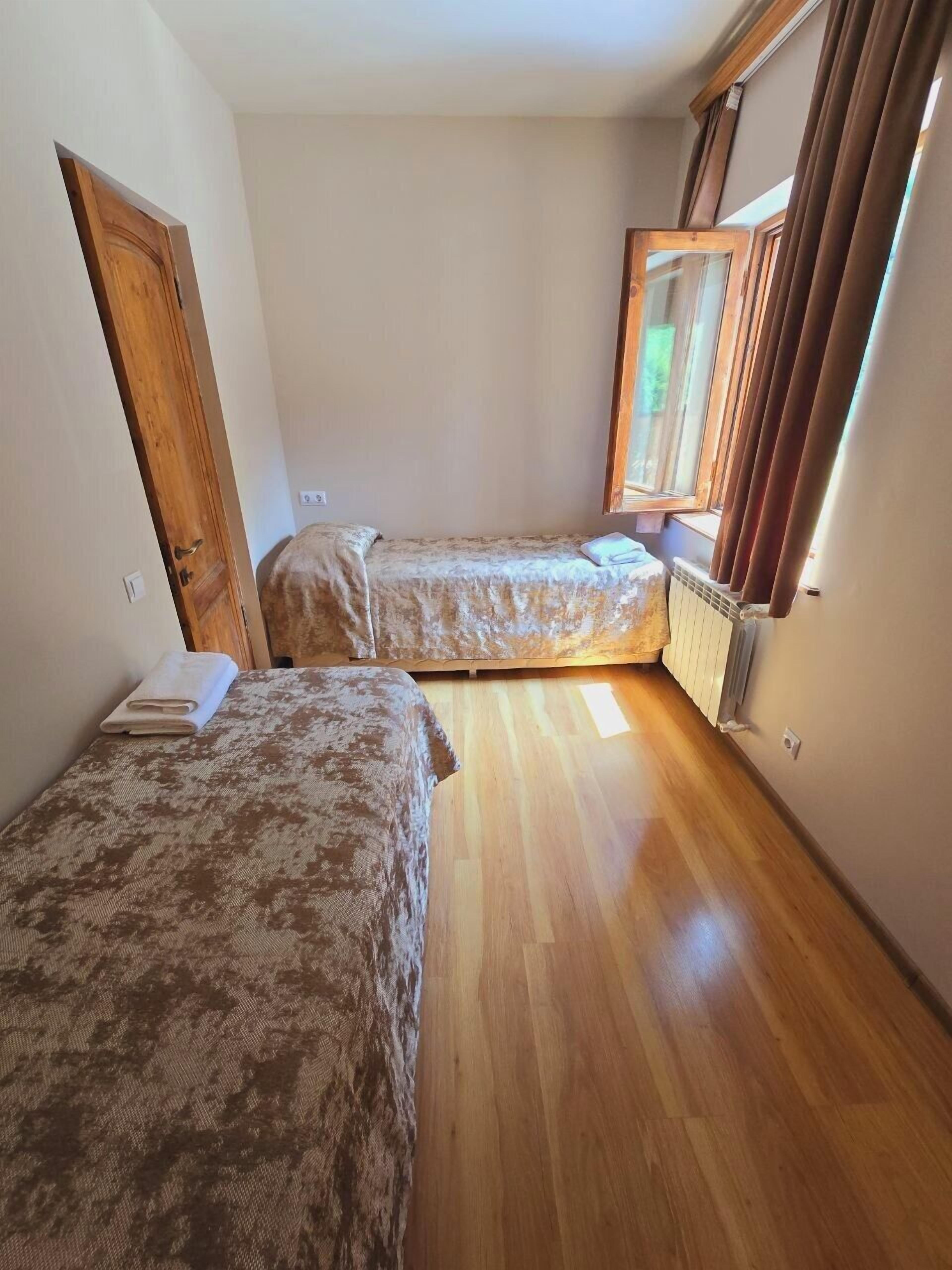 A room with two beds, wooden flooring, a window with curtains, and a radiator.