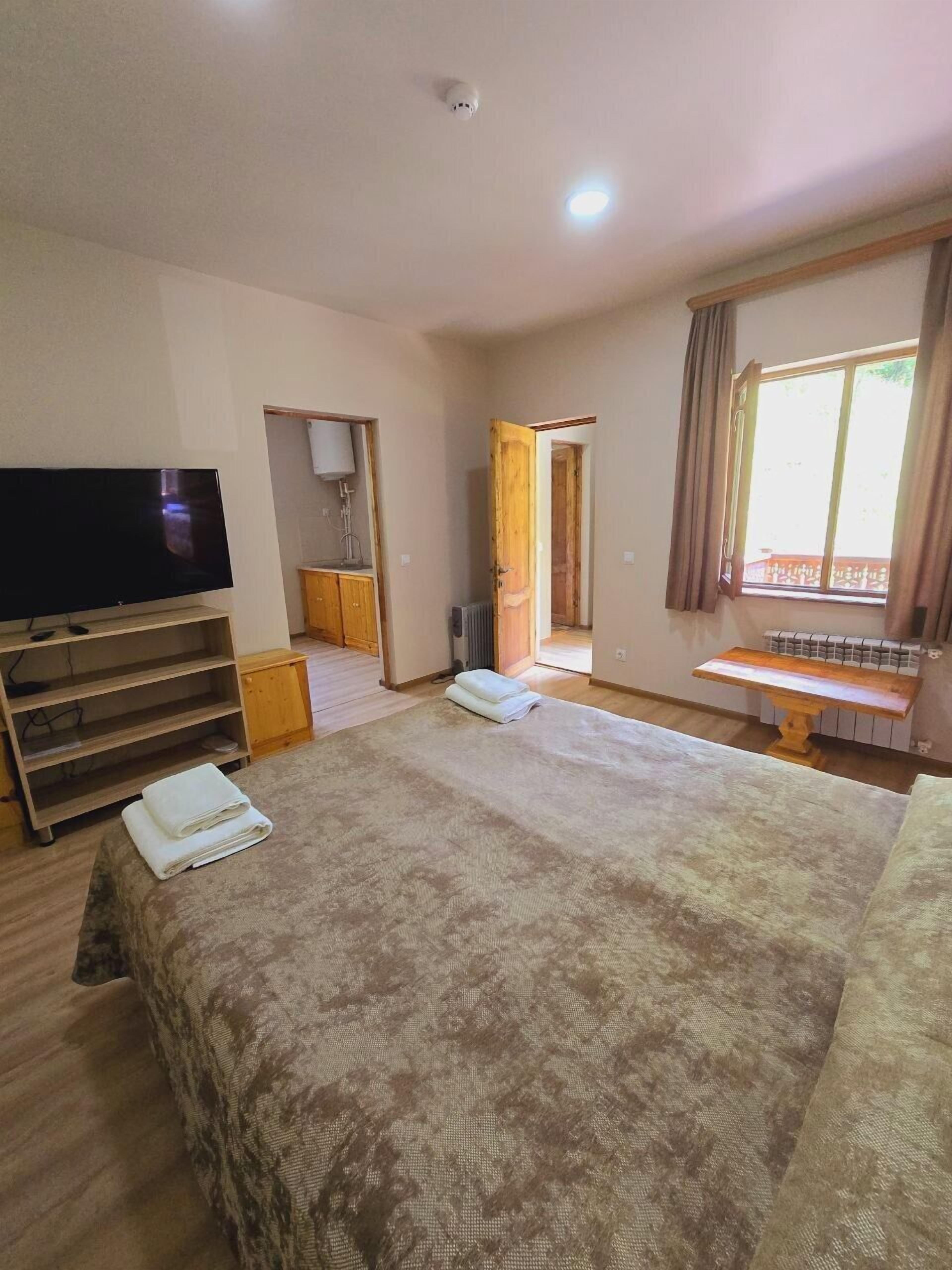 A bedroom with a large bed, a TV on a stand, a wooden cabinet, and a window with curtains.