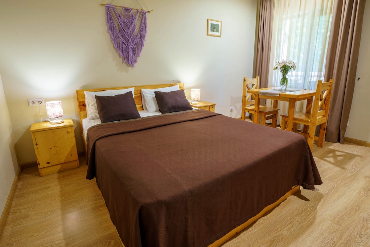 Een slaapkamer met een groot bed, houten hoofdbord, nachtkastjes met lampen, een kleine tafel met stoelen en een paars macramé wanddecoratie.