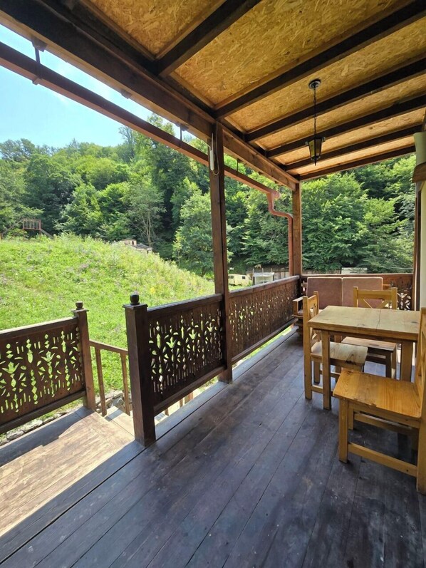 A covered wooden deck with a table and chairs, surrounded by a wooden railing, overlooking a lush green landscape.