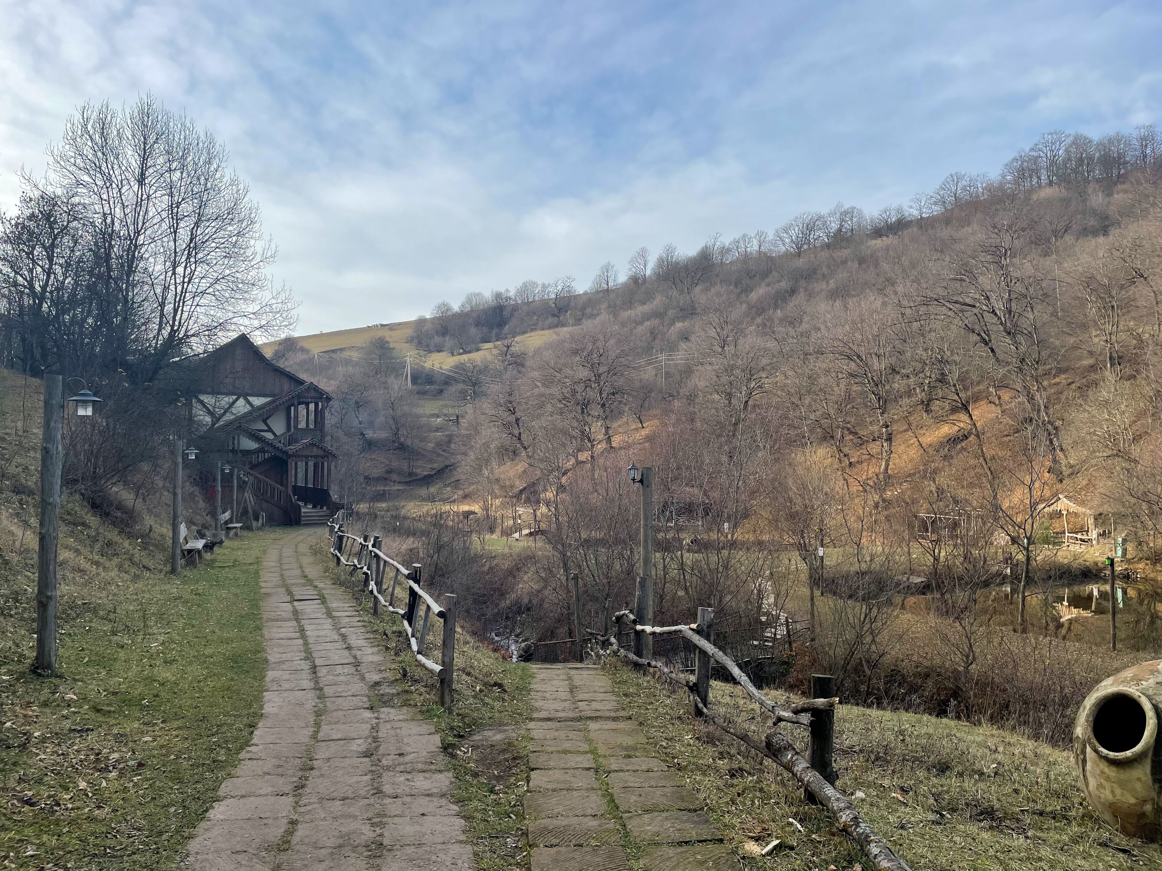 A stone pathway leading to a traditional wooden building with a balcony, surrounded by a rural landscape with hills and sparse trees.