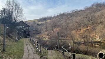 A stone pathway leading to a traditional wooden building with a balcony, surrounded by a rural landscape with hills and sparse trees.
