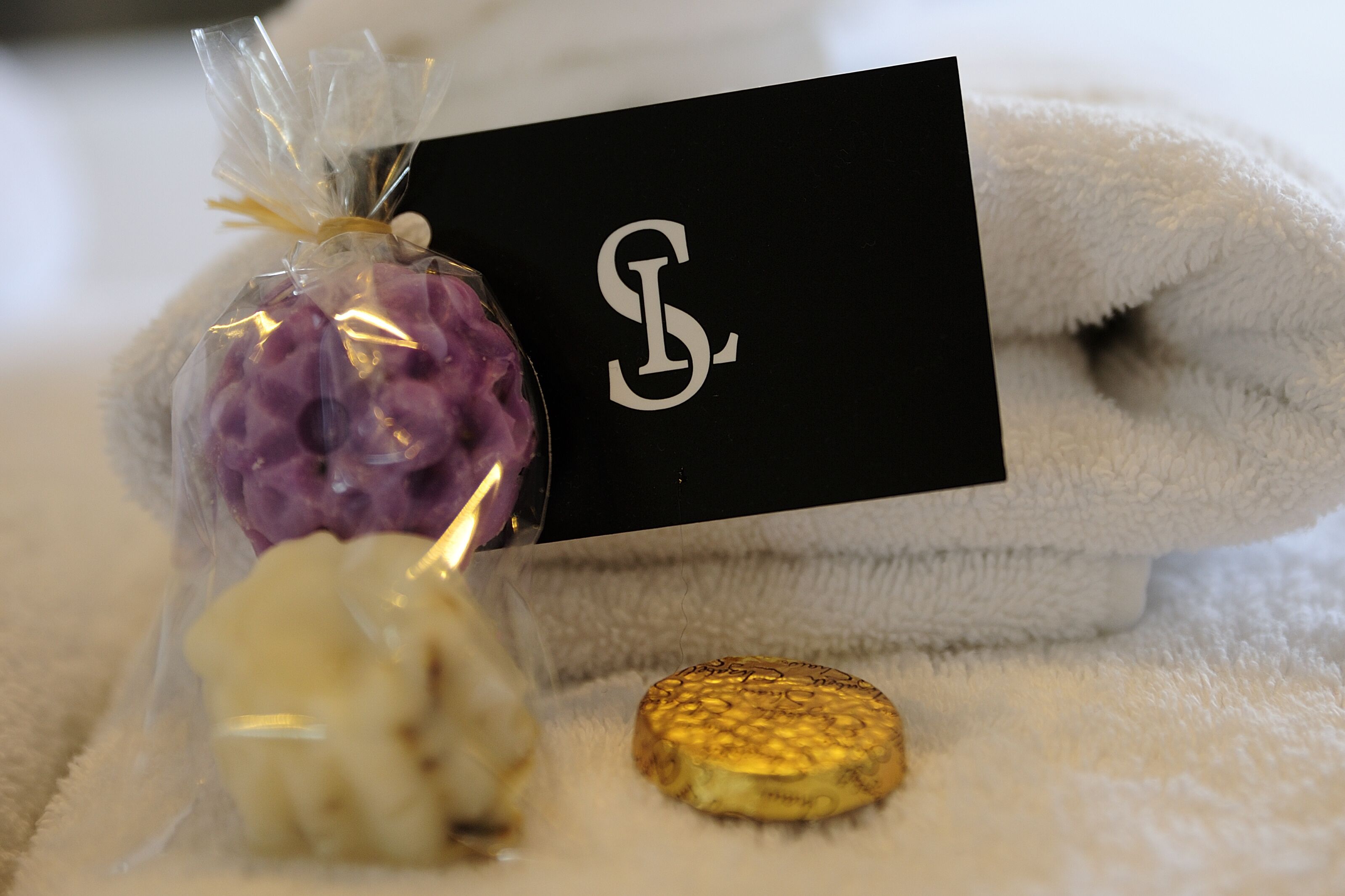 Um cartão preto com uma monógrama branca, uma toalha branca e fofa e um objeto dourado pequeno sobre uma superfície branca e fofa.
