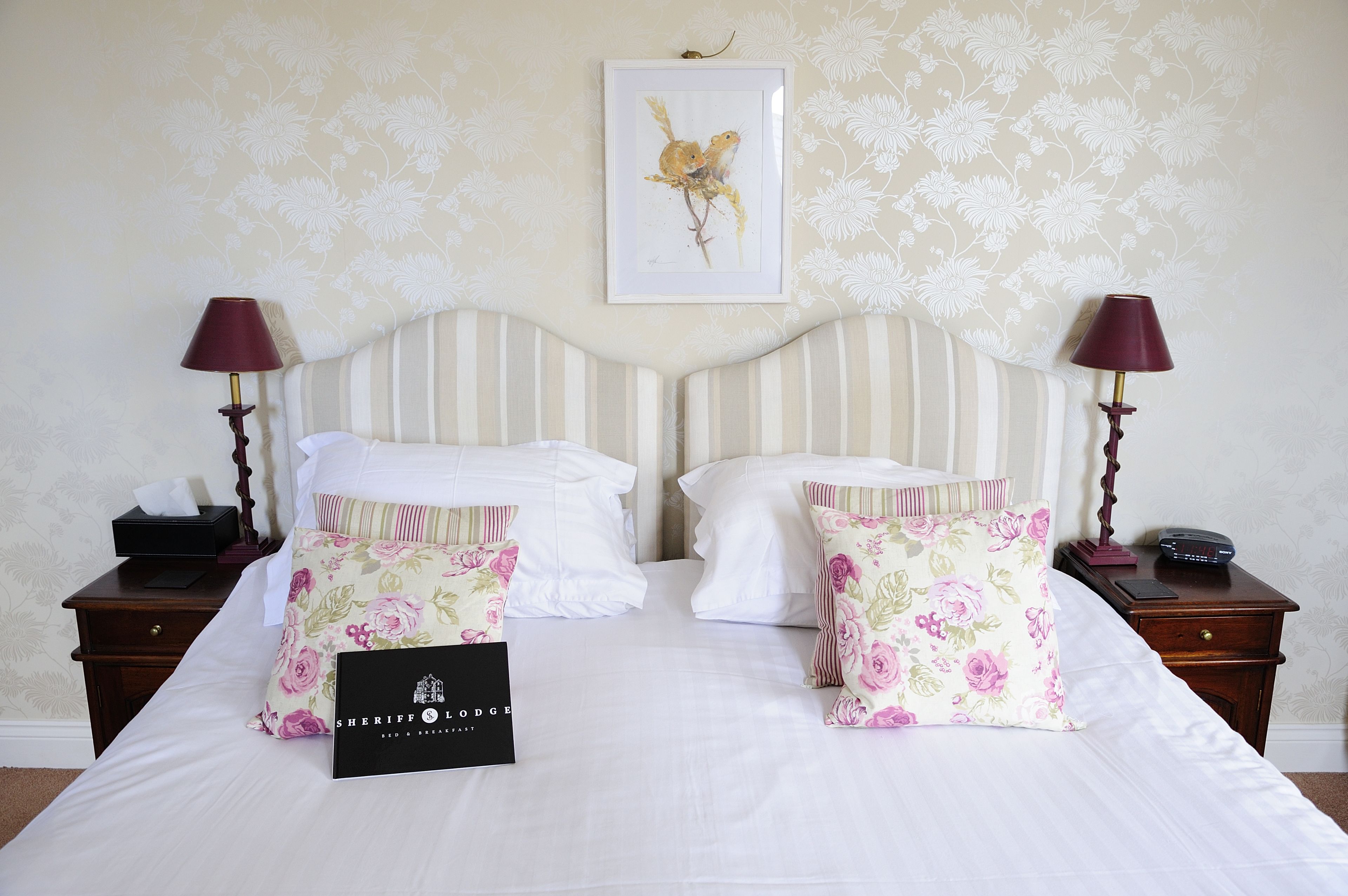 A neatly made double bed with floral pillows, a framed artwork above, and a small bedside table with a lamp and a phone.