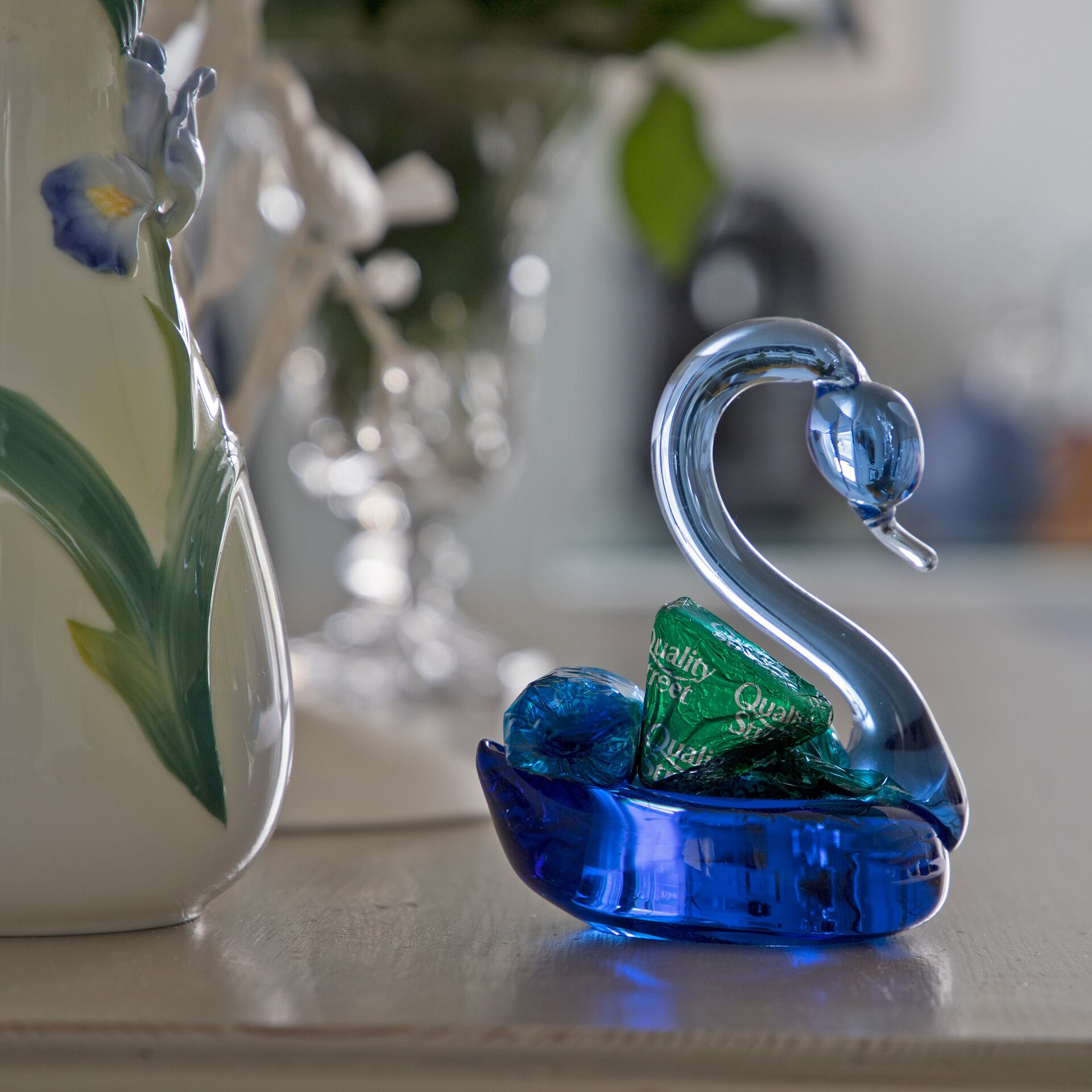 A swan-shaped glass ornament with a green and blue design, placed on a surface next to a clear glass vase with a floral pattern.