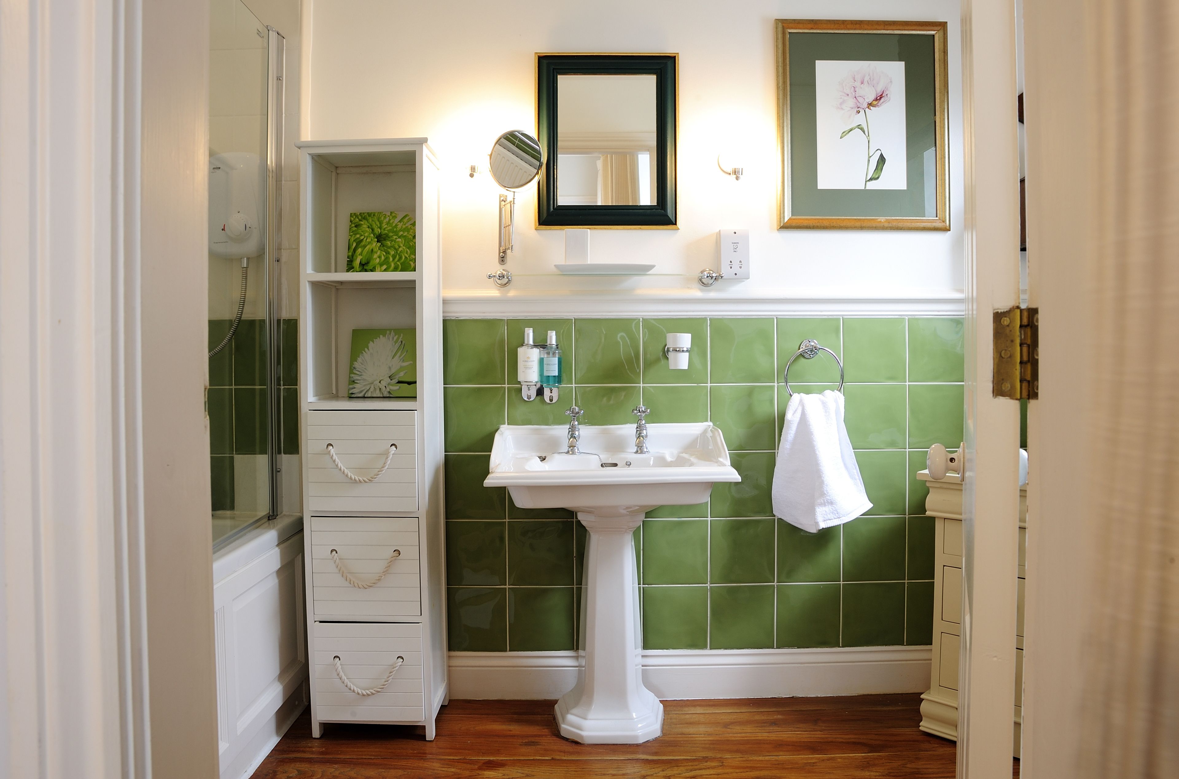 A bathroom with a pedestal sink, green tiled walls, a white vanity with drawers, a framed mirror, and a framed picture on the wall.
