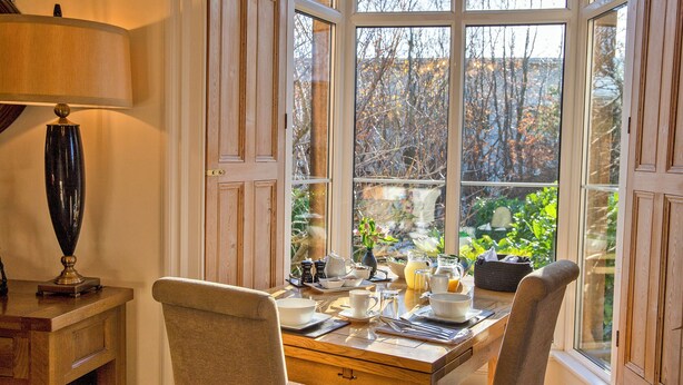 A dining area with a wooden table set for two, a lamp, and a view of trees through large windows. - Sheriff Lodge (Matlock)