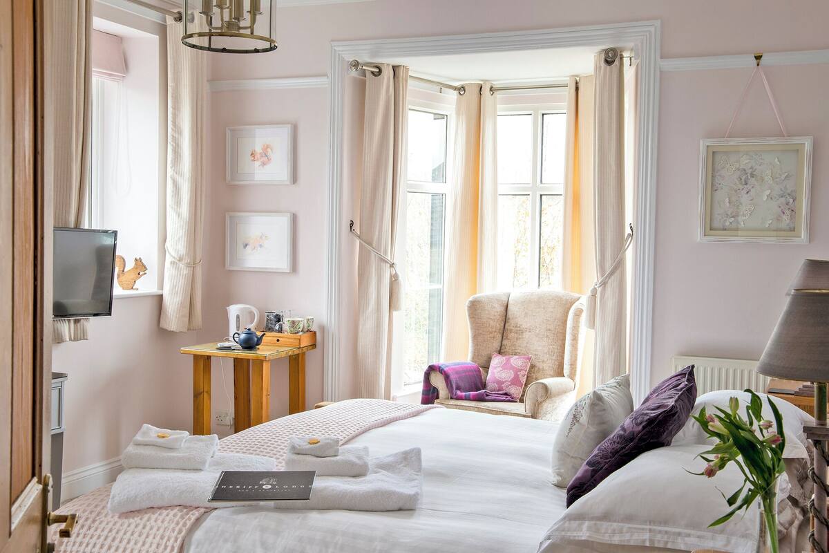 A hotel room with a bed, a chair, a small table with a kettle, and a TV mounted on the wall.