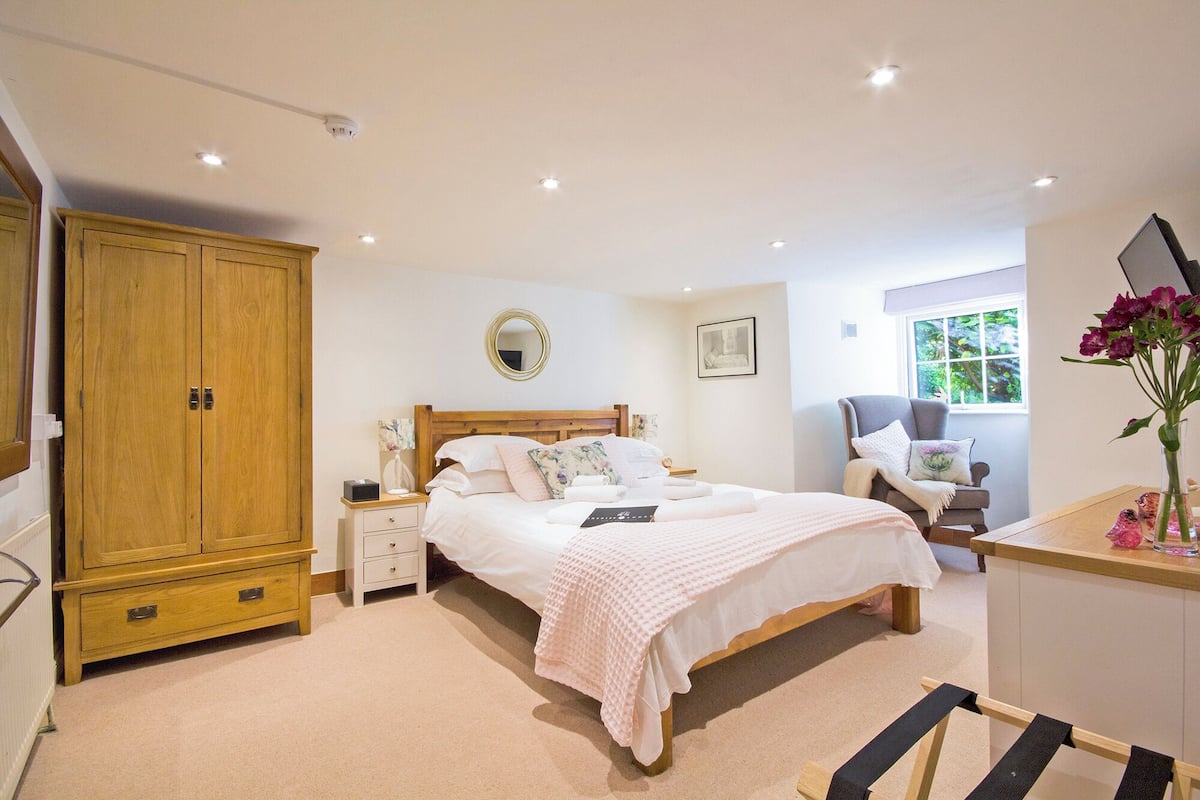 A bedroom with a wooden wardrobe, a bed with white bedding, a bedside table, a chair, a window with a view, and a vase with flowers.