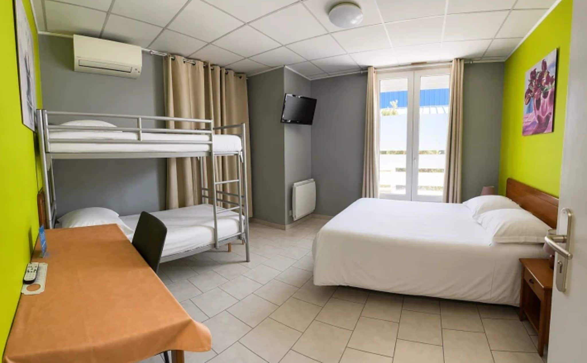 A hotel room with bunk beds, a single bed, a TV, a table with a chair, and a window with curtains.