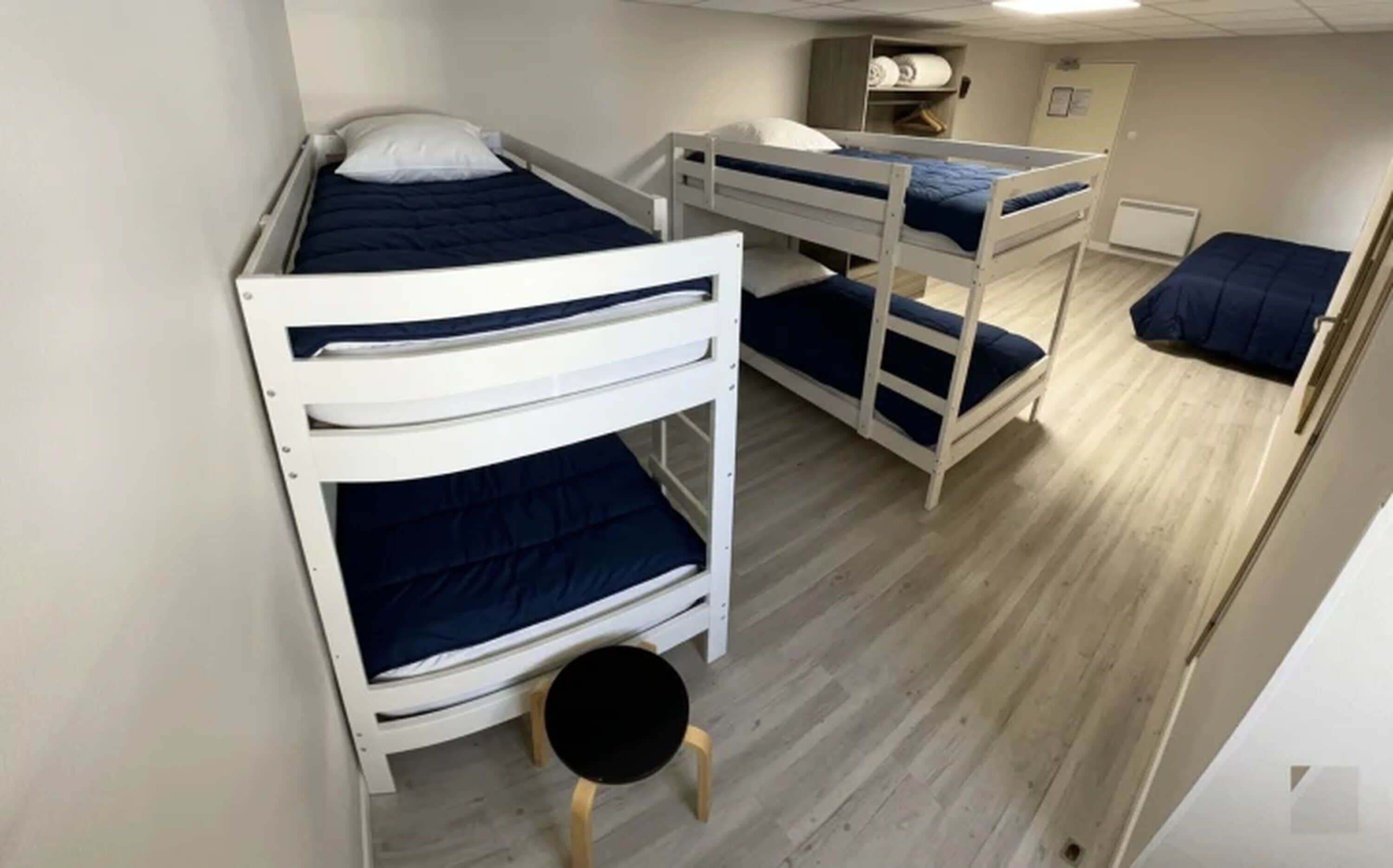 Habitación con literas, un taburete redondo, suelo de madera y una pequeña estantería con objetos.