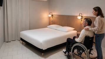 A bedroom with a bed, two wall-mounted lights, a window with curtains, and a wheelchair.